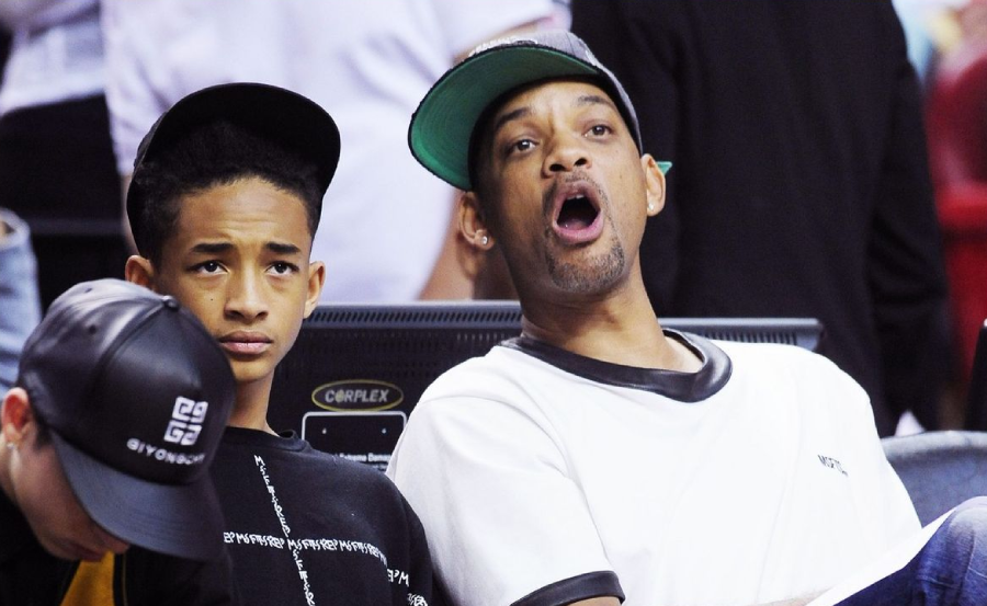 A boy and a man sit side by side at a sports event. The boy looks serious while the man next to him, wearing a cap and white shirt, has his mouth open in surprise or excitement.