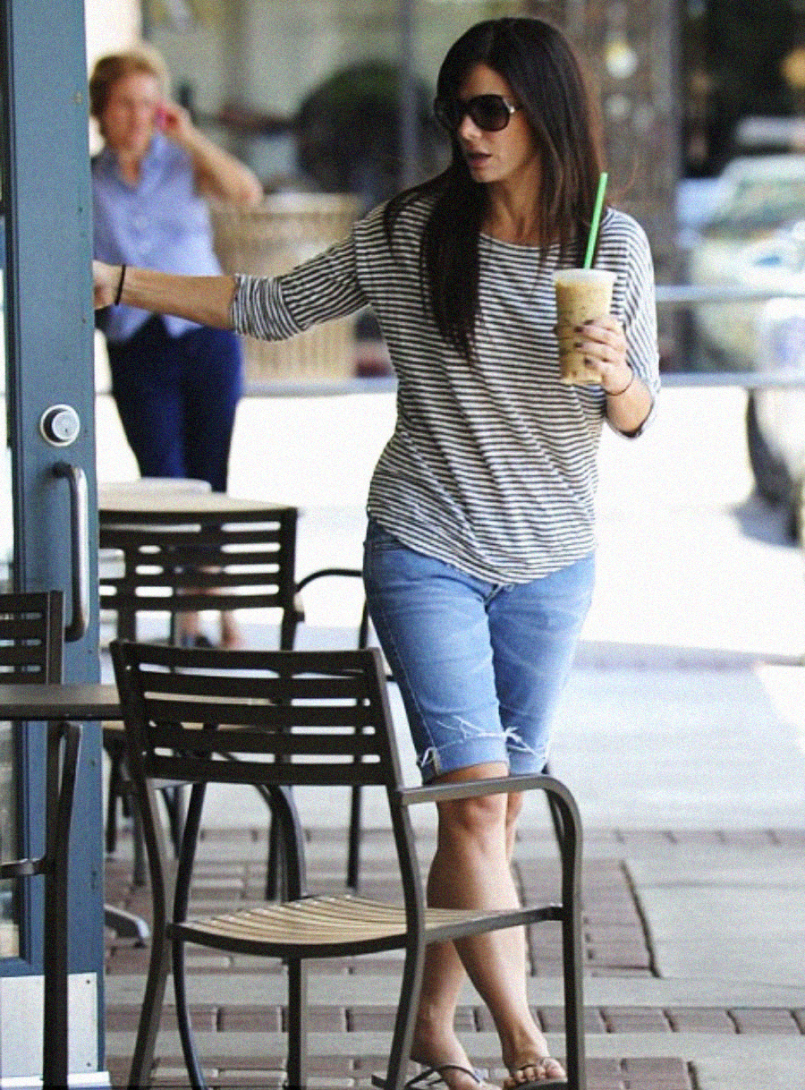 A woman wearing sunglasses, a striped shirt, and knee-length denim shorts holds an iced coffee and opens a glass door outside a cafe, with another person walking in the background.