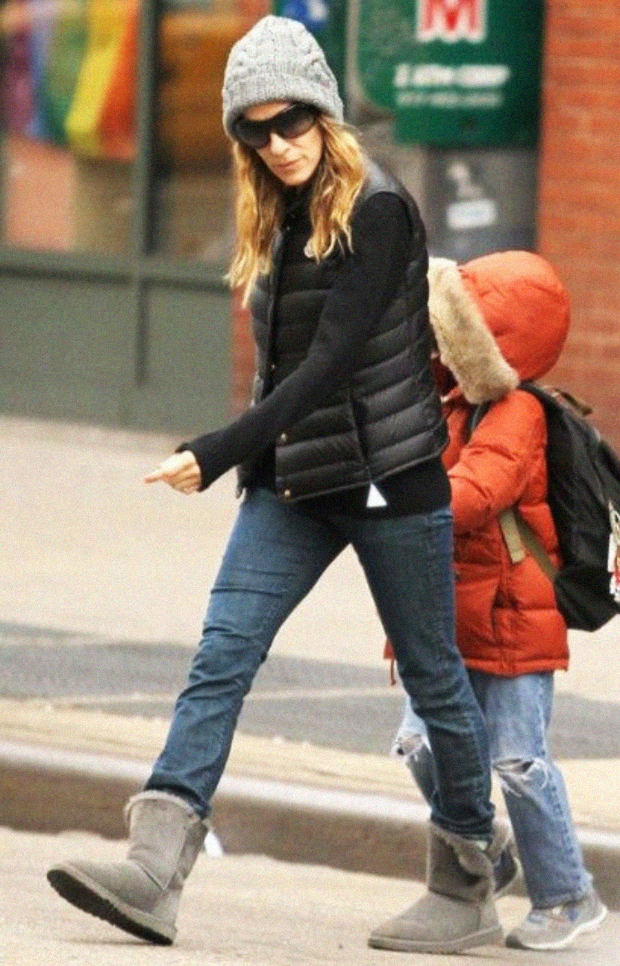A woman in a gray beanie, sunglasses, black puffer vest, jeans, and gray boots walks beside a child in a red coat with a fur-lined hood and a backpack on a city sidewalk.