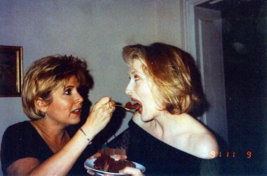 Two women are indoors; one is feeding the other a bite of cake with a fork. The woman being fed has her mouth open wide and is wearing an off-shoulder black top. There is a plate with cake in the scene.