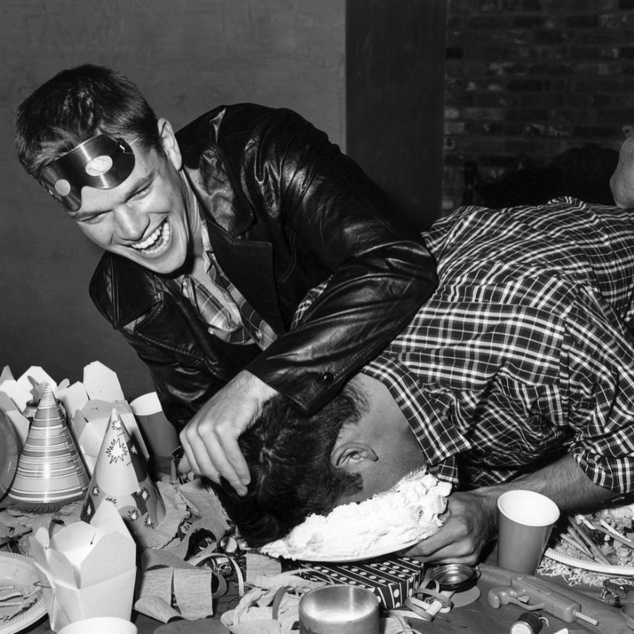 Two young men at a party, one playfully pushes the other's face into a cake on a table covered with party hats, cups, and decorations. Both are laughing. The scene is in black and white.