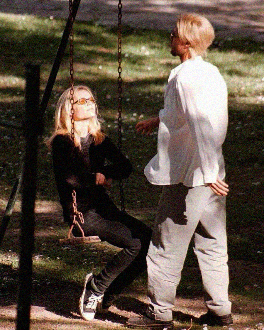 A woman wearing sunglasses sits on a swing, leaning back with her arms crossed, while a man in a white shirt and gray pants stands nearby, hands behind his back. They appear to be outdoors in a park.