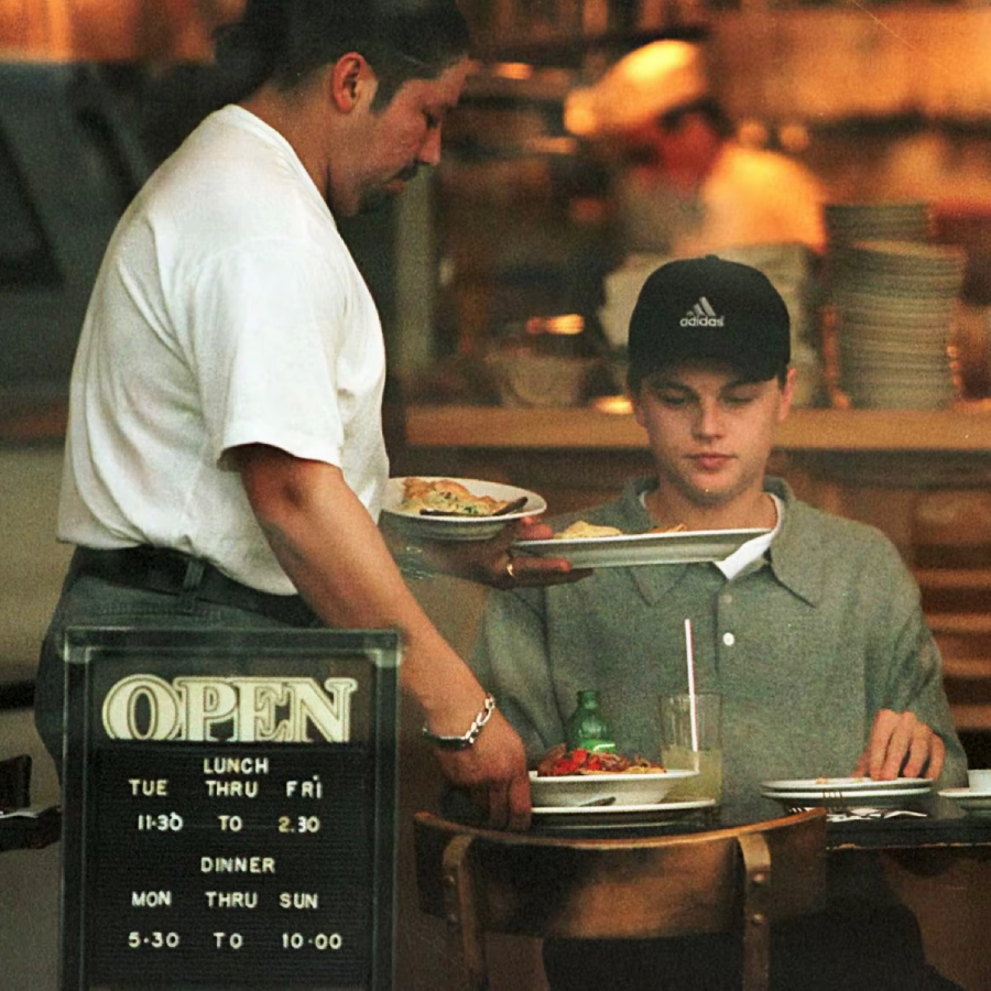 A server places plates of food in front of a young man sitting at a restaurant table. A glass of water and a salad are on the table. An "OPEN" sign with business hours is visible in the window.