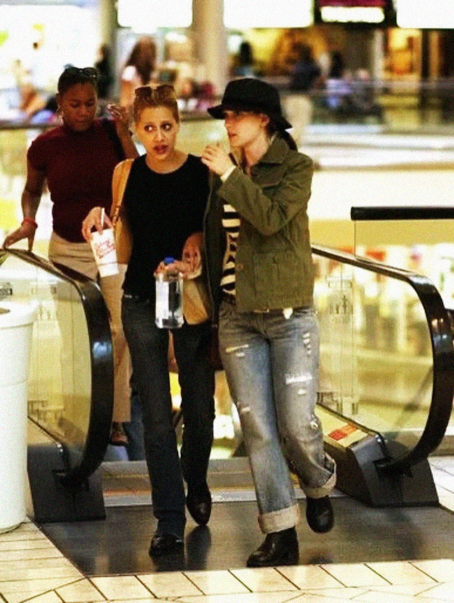 Two women walk side by side up an escalator in a shopping mall, one wearing a black tank top and holding a drink, the other in a green jacket and black hat. A third woman is seen behind them.