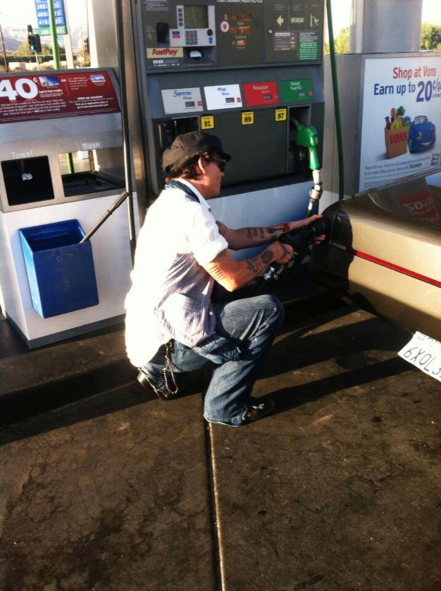 A person wearing a hat, sunglasses, and jeans crouches while pumping gas into a car at a gas station on a sunny day. Gas pumps, a blue trash bin, and a sign advertising discounts are visible.