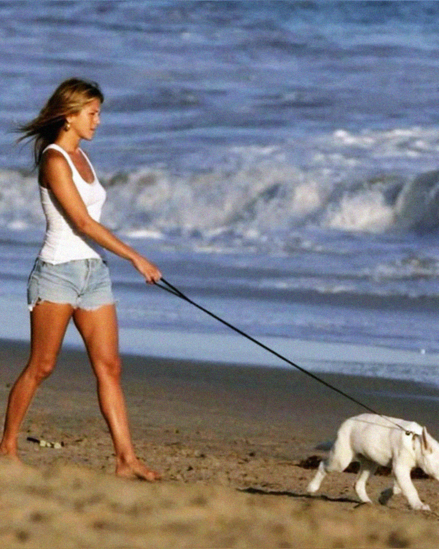 A woman in a white tank top and denim shorts walks a small dog on a leash along a sandy beach with waves in the background.