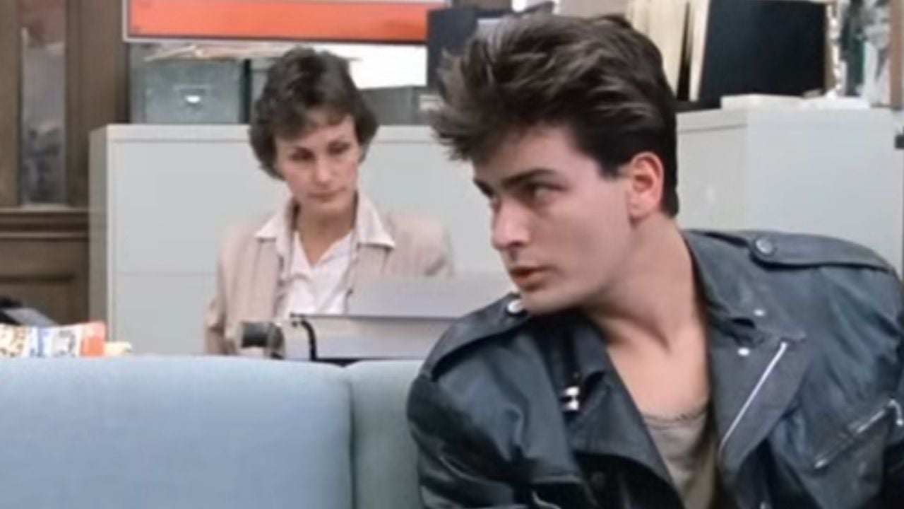 A young man in a leather jacket sits on a couch, looking to the side. In the background, a woman with short hair sits at a desk, looking down at papers.