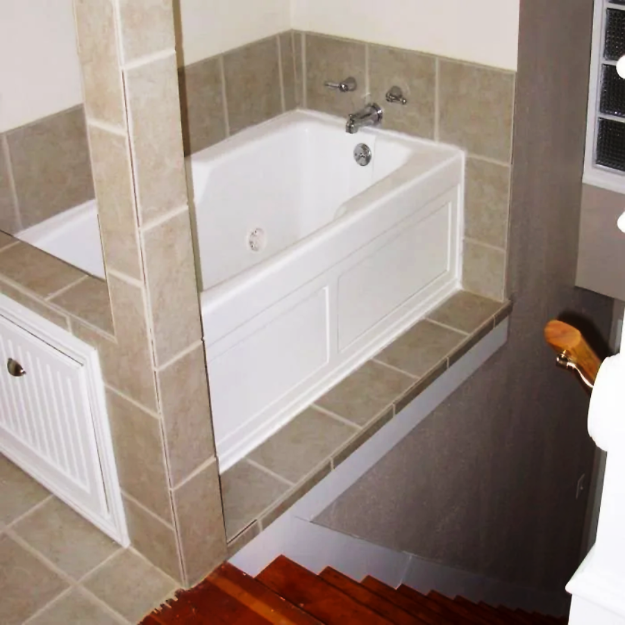 A white bathtub with beige tile surround is positioned dangerously close to the edge of a staircase, with no barrier between the tub and the open stairwell below.