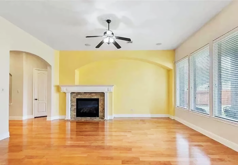 A spacious living room with light wood floors, a yellow accent wall, a white fireplace, ceiling fan with lights, arched doorways, and large windows letting in natural light.