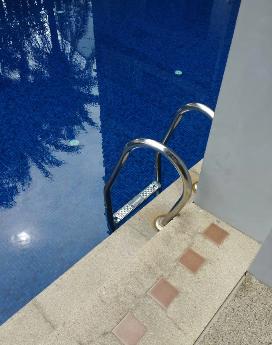 A swimming pool ladder descends into clear blue water beside a tiled pool deck. The water reflects palm trees, and the pool edge is lined with beige and brown square tiles.