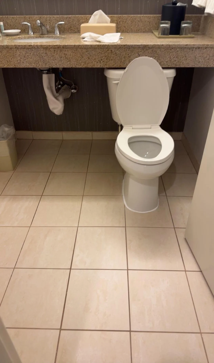 A clean bathroom with a white toilet, an open lid, beige tiled floor, a granite countertop above, and a trash can to the left. Towels, toiletries, and tissue boxes are on the countertop.