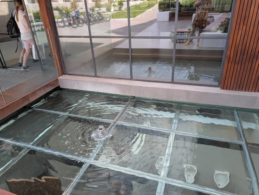 A glass floor covers a shallow pool with a bubbling water feature underneath. Reflections of two people and outdoor scenery are visible in the glass and the window.