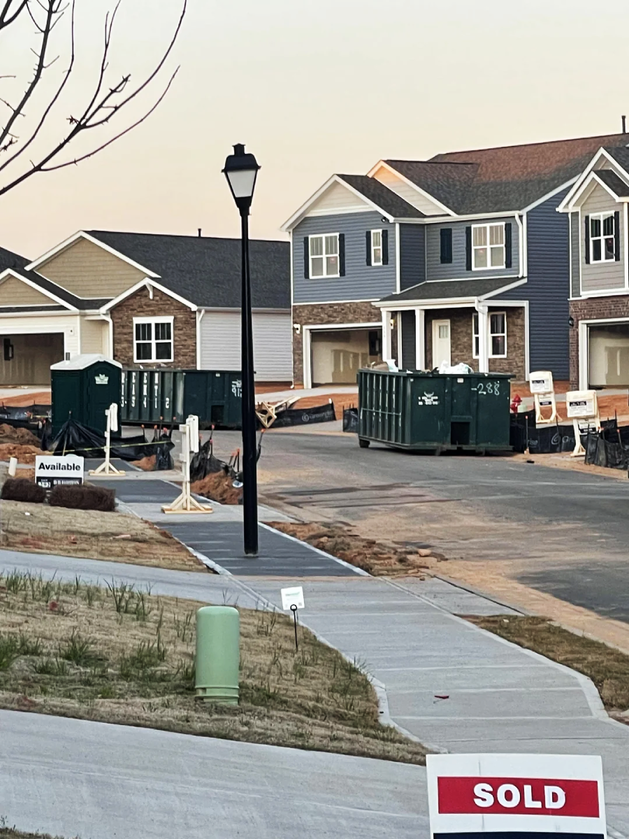 Newly built suburban houses line a street with construction dumpsters in driveways. A "SOLD" sign is in the foreground, and sidewalks curve through the neighborhood under a streetlight.