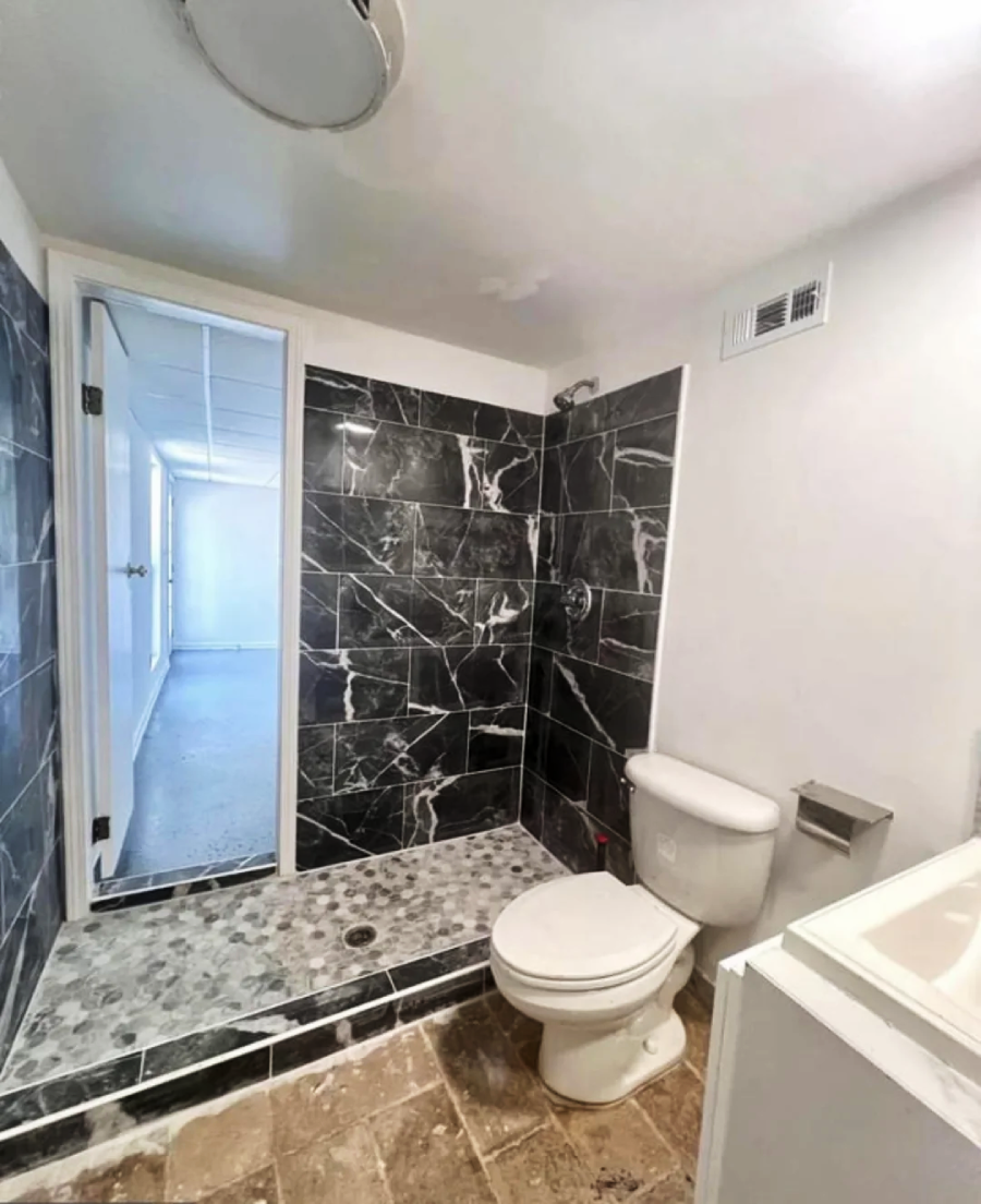A bathroom with a toilet positioned inside a shower area with black marble tiles, next to a small sink; the floor transitions from tan tiles to black marble in the shower.