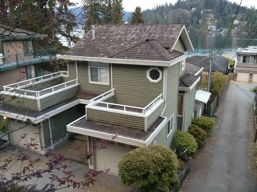 Two-story green house with white trim, multiple balconies, and a garage, situated on a sloped street with bushes in front and a lake and forested hills visible in the background.