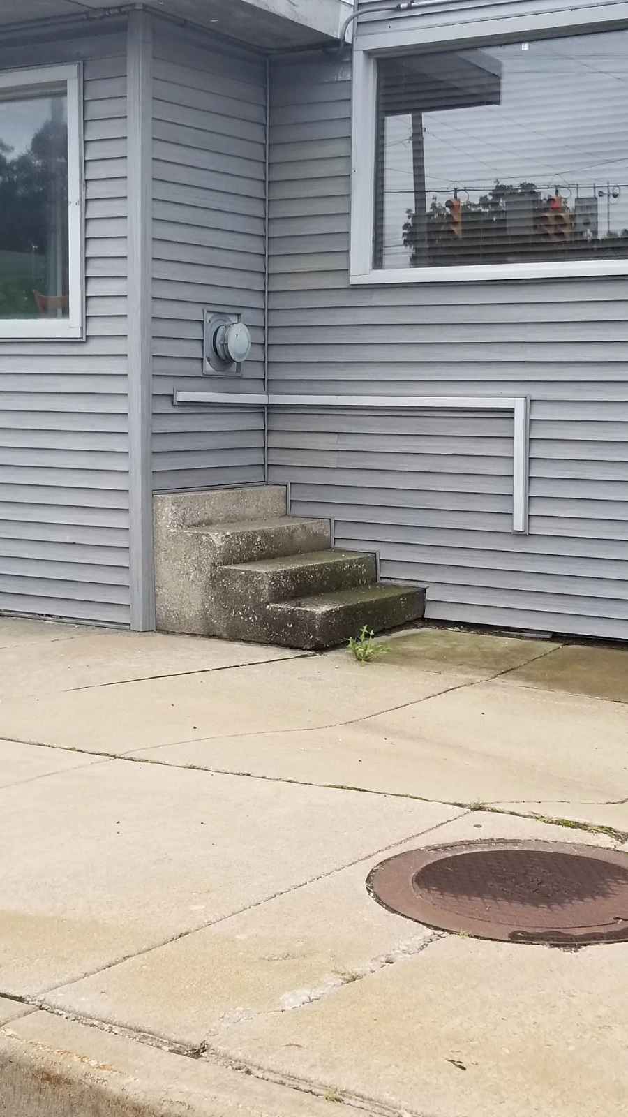 A set of concrete steps leads directly into the siding of a gray building, with no door or entryway, next to a sidewalk and a manhole cover. Grass grows in a crack near the bottom step.