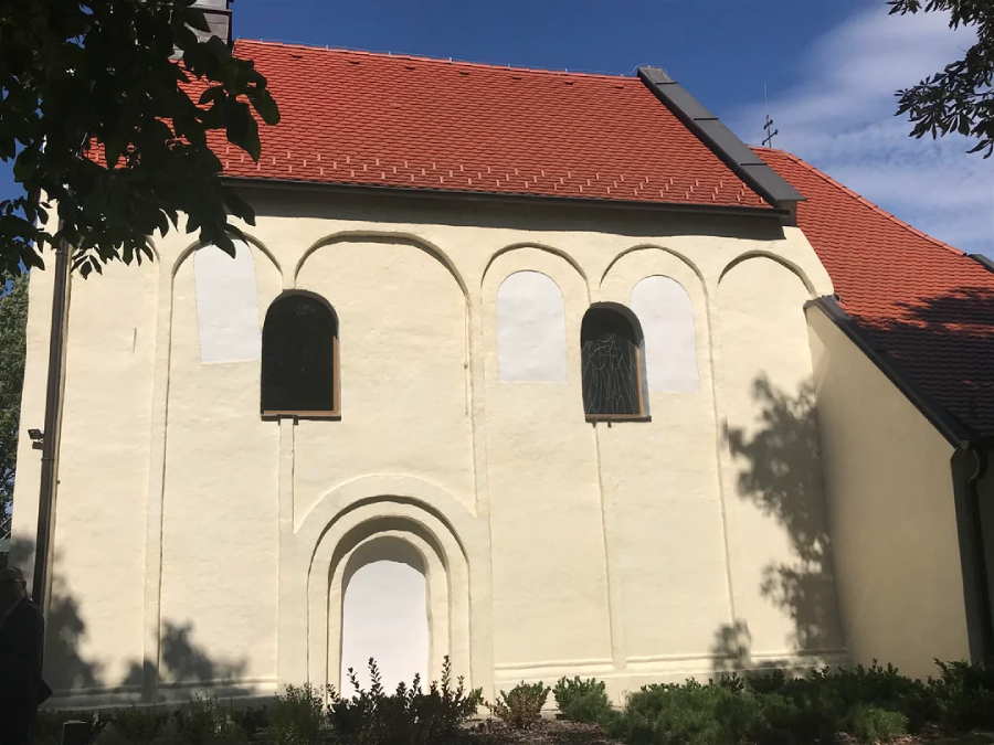 A cream-colored church with a red tiled roof, arched windows, and an arched doorway, partially shaded by trees. The building features decorative arches on its exterior wall and simple landscaping in the foreground.