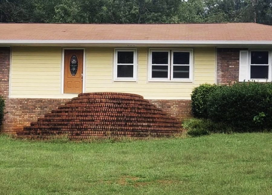 A house with a yellow and brick exterior has a front door accessed by a large, dome-shaped mound of brick steps, but no railings or clear entryway path, surrounded by grass and shrubs.