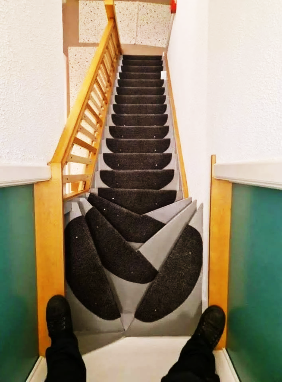 A staircase with dark, circular-patterned carpet and an optical illusion at the bottom that makes the steps appear twisted and warped; a person’s feet are visible at the top of the stairs.