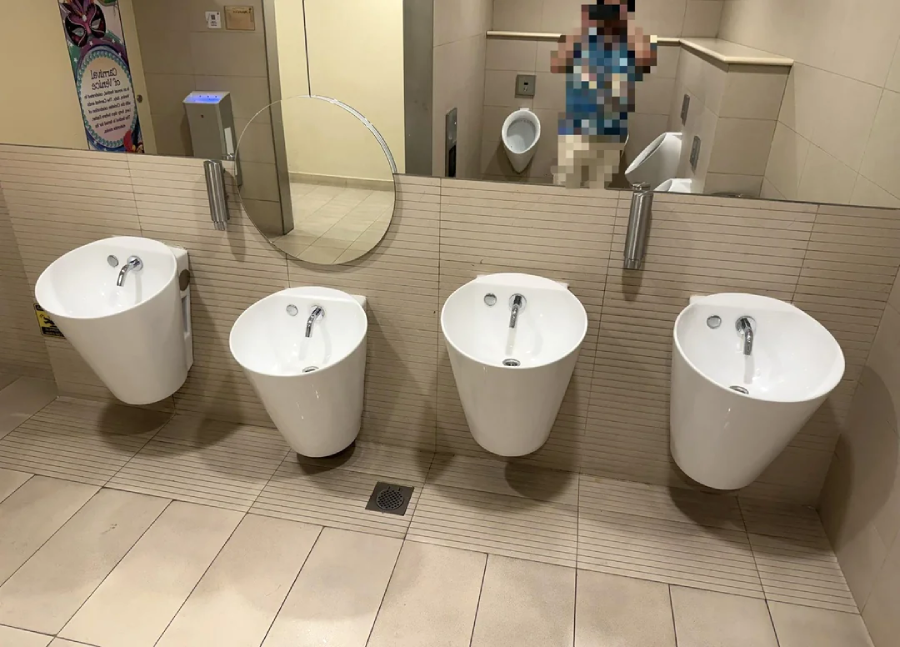 Four white sinks of varying heights are mounted on a tiled wall in a public restroom, with a round mirror above them. A blurred person is reflected in the mirror.