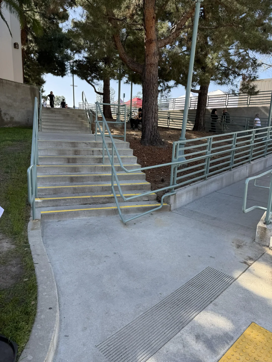 A wide concrete staircase and parallel wheelchair ramp with green railings are outdoors, surrounded by tall pine trees and a fence. Yellow tactile paving is visible on the sidewalk in the foreground.