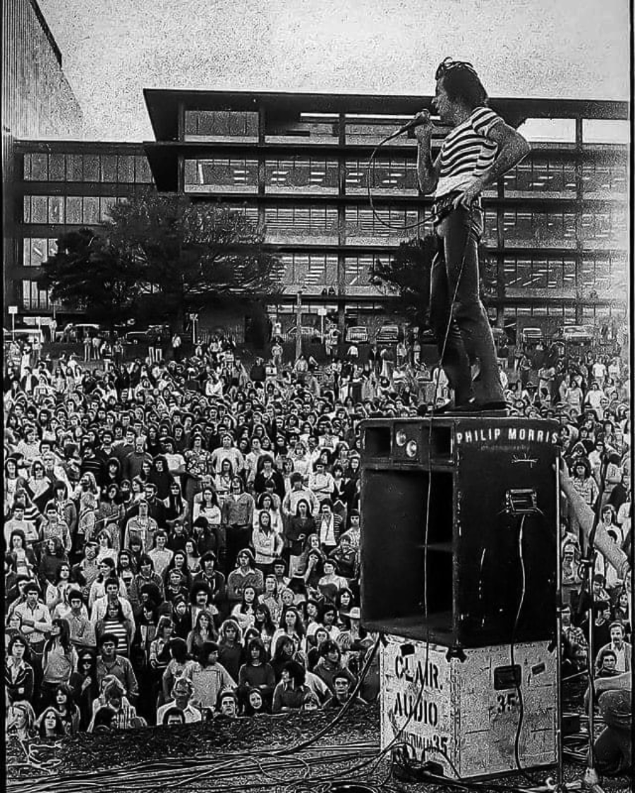 A singer stands on top of a speaker, holding a microphone and performing to a large crowd gathered outdoors in front of a modern multi-story building. The scene appears to be from a past decade.