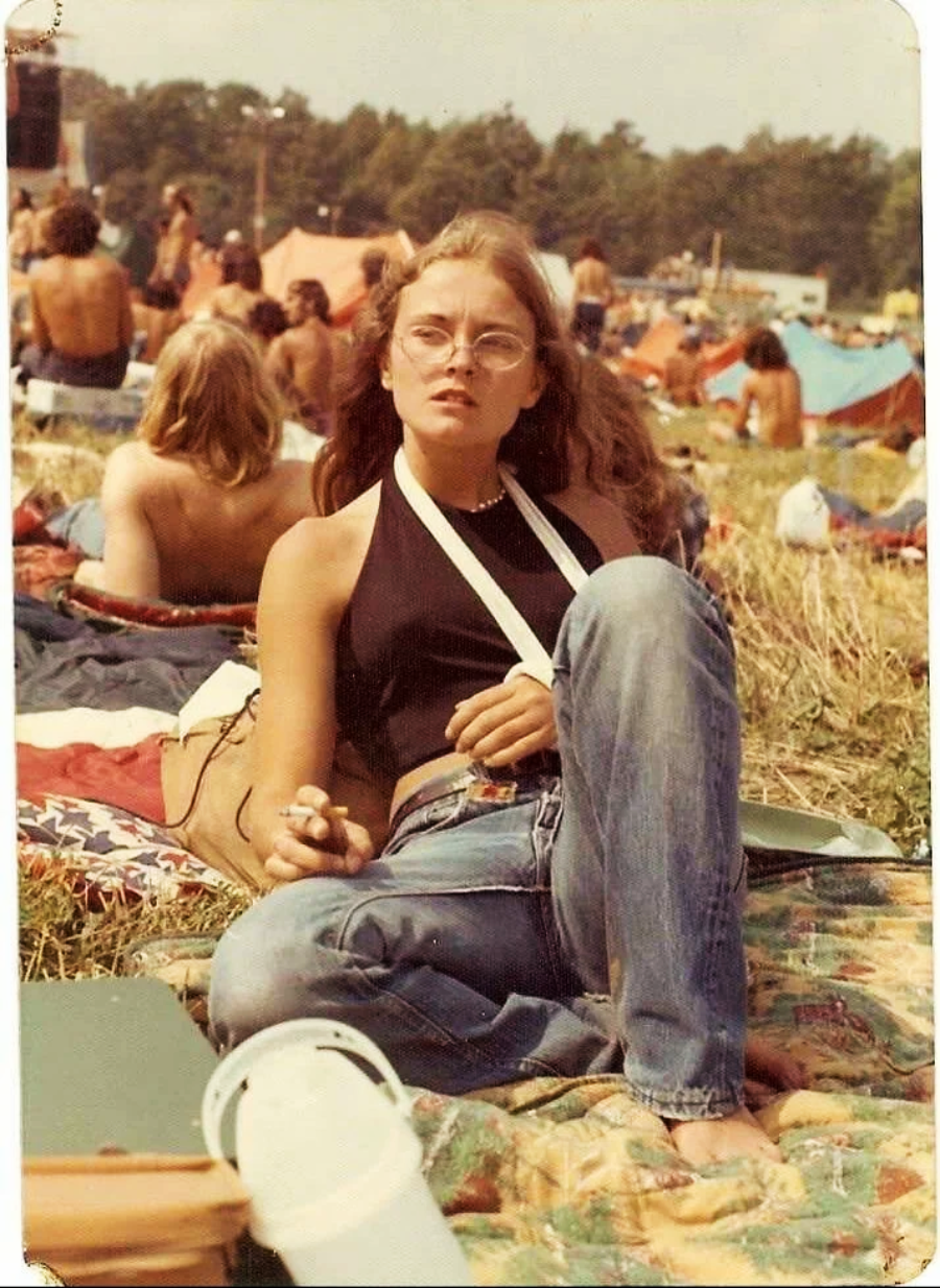 A young woman with long hair and glasses sits cross-legged on a blanket at an outdoor event. She wears a black halter top, blue jeans, and has a cigarette in hand. People relax on the grass and tents are visible in the background.