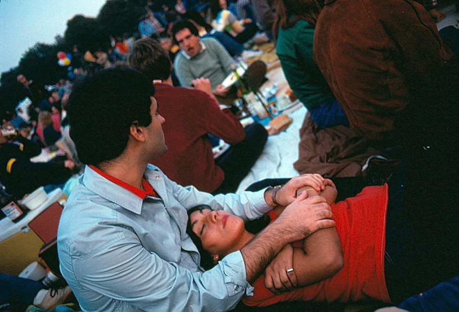 A man sits on a picnic blanket, gently holding a woman who is lying with her head in his lap. They are surrounded by other people sitting closely together outdoors, suggesting a relaxed, social gathering.
