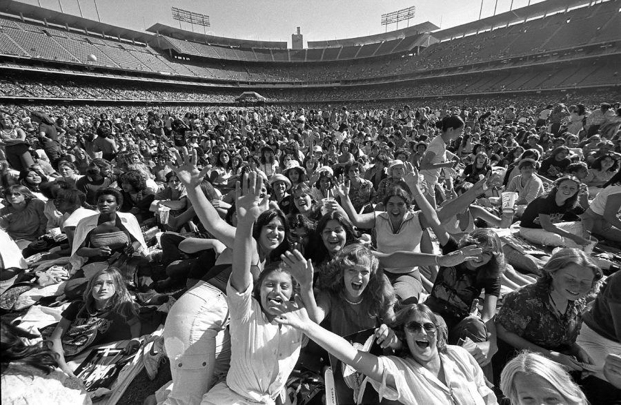 A large, excited crowd of mostly young people sits and waves at the camera inside a stadium during a daytime event; many are smiling, cheering, and reaching out enthusiastically.