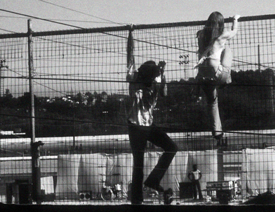 Two people climb a wire fence outdoors, with one near the top and the other halfway up. In the background, there are more people, vehicles, and structures visible under a clear sky.
