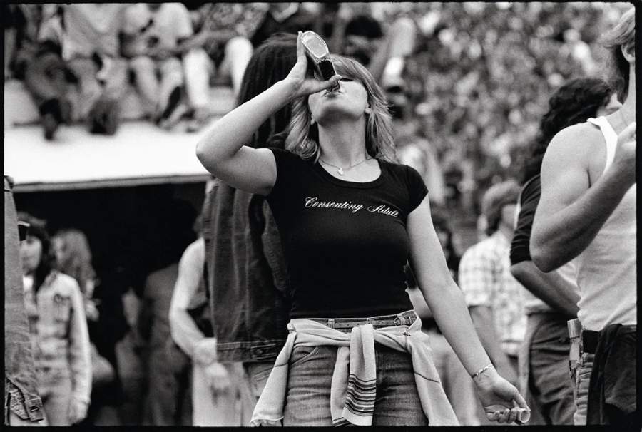 A woman in a "Consulting Adult" t-shirt drinks from a bottle at an outdoor event, with people and trees in the background. The photo is black and white, capturing a casual, lively atmosphere.