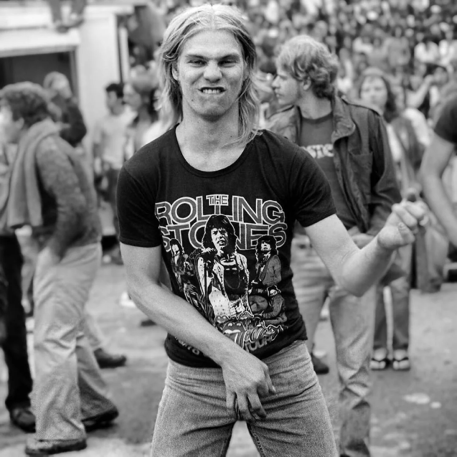 Young man with long hair enthusiastically plays air guitar, wearing a Rolling Stones t-shirt and jeans, at an outdoor event with a crowd of people in the background. The image is in black and white.
