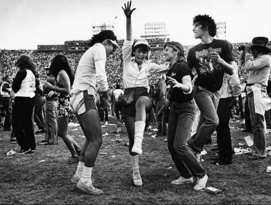 Four young people dance energetically on a grassy field at an outdoor event, surrounded by a large crowd of spectators in the background. The atmosphere appears joyful and lively.