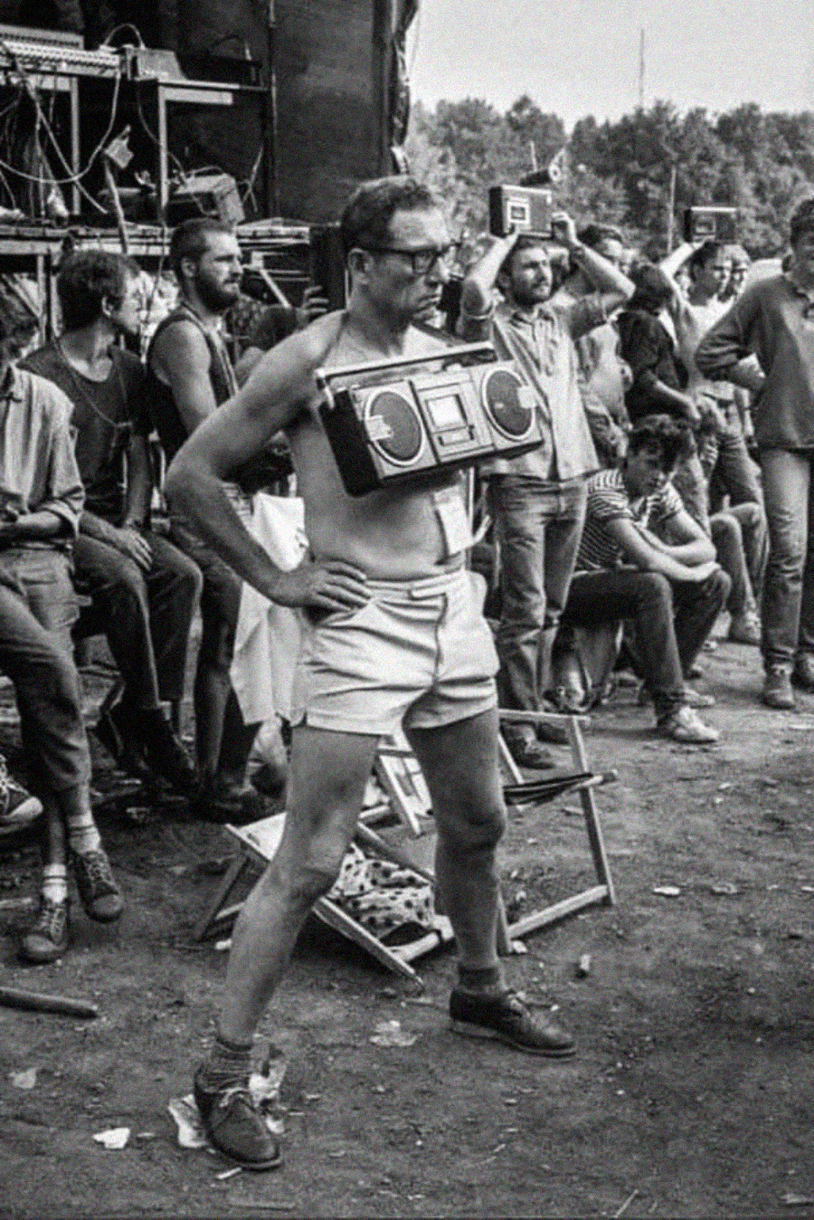 A man in shorts and glasses stands confidently with a boombox strapped to his chest at an outdoor event, surrounded by a crowd of casually dressed people. The scene appears lively and spirited.