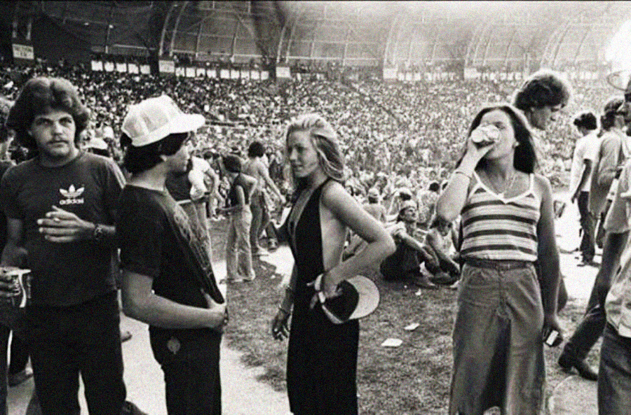 A black-and-white photo of a crowded outdoor concert or festival in the 1970s; several young adults stand in the foreground, talking and drinking, while a packed stadium crowd fills the background.