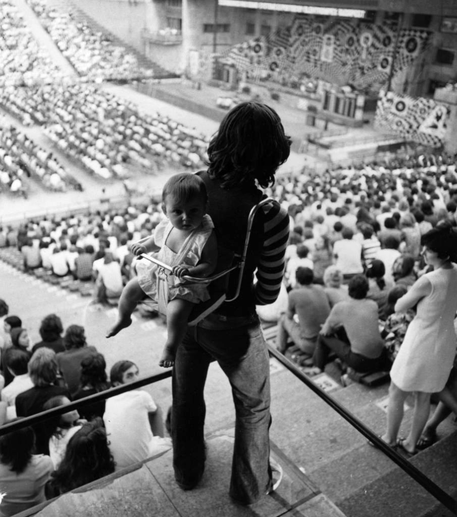 A person with long hair stands facing a crowded outdoor concert stage, carrying a baby in a front carrier. The audience sits on stone steps, watching the performance.