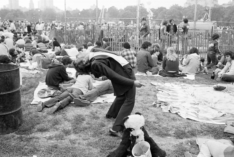 A person in a suit and sash leans backward dramatically in a crowded park. People sit on blankets and grass, with a fenced area and buildings in the background. A stuffed panda and a bucket are in the foreground.