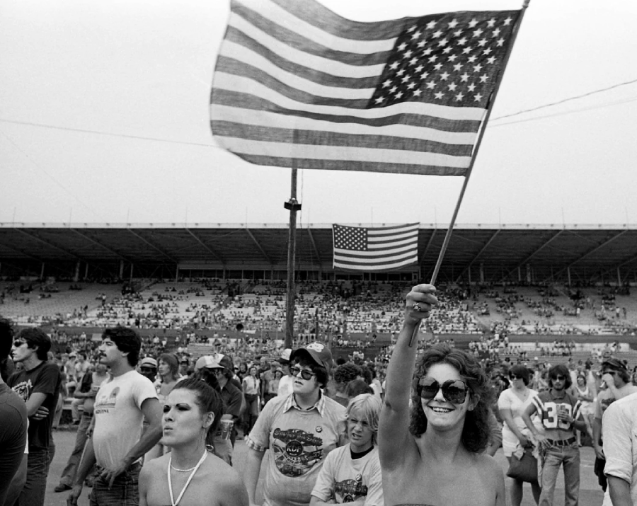 A woman in sunglasses smiles and waves a large American flag at an outdoor stadium event, with a crowd of people around her and another American flag hanging in the background.
