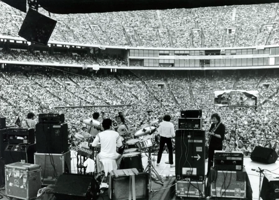 Black-and-white photo of a band performing on stage in a large stadium, viewed from behind the musicians, with a vast audience filling the seats and stands.