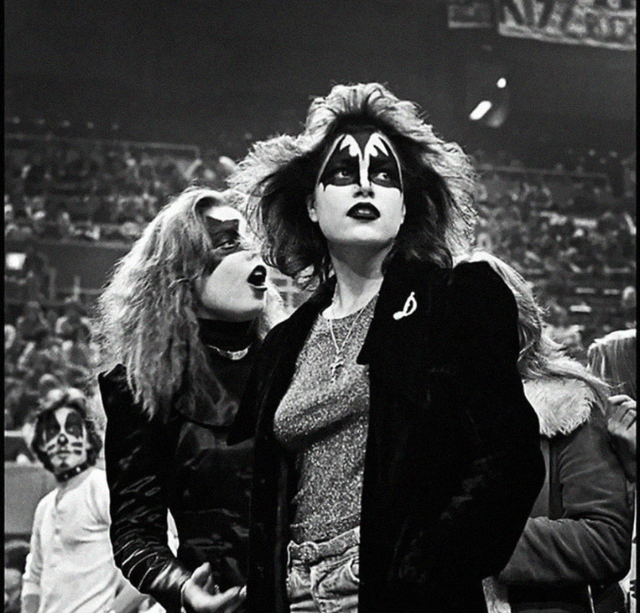 Two women in a crowded arena wear black leather jackets and dramatic face paint similar to the rock band KISS, standing among other fans, all appearing excited and energetic.
