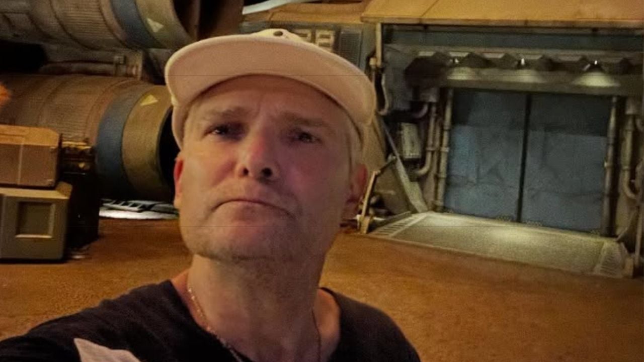 A man in a white cap takes a selfie in front of a sci-fi style building with industrial pipes and large machinery.