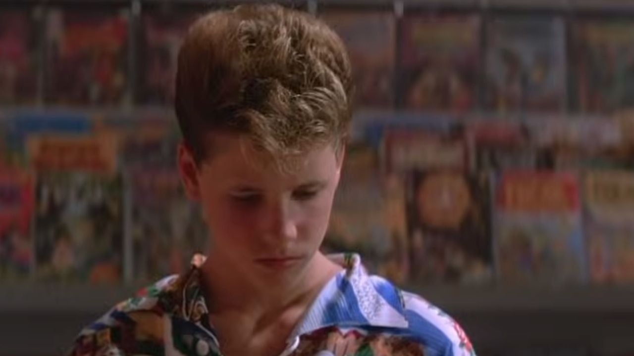 A boy with short, curly hair wearing a colorful shirt looks down, standing in front of a wall filled with comic books.