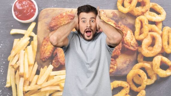 A surprised man in a gray t-shirt holds his hands to his head, standing in front of a background featuring fries, fried chicken, onion rings, and a cup of ketchup.
