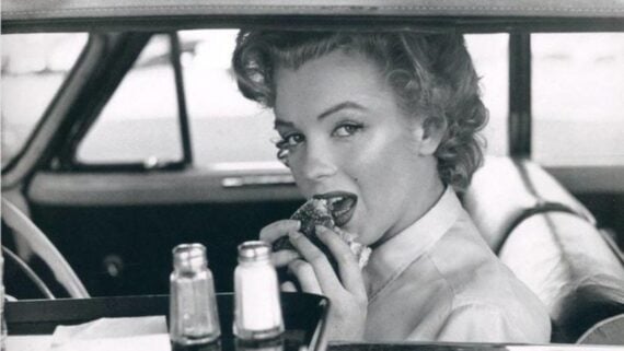 A woman with styled blonde hair sits in a car and looks at the camera while eating a burger; salt and pepper shakers and a food tray are visible in the foreground.