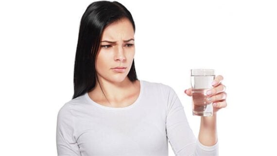 A woman with long dark hair in a white shirt looks at a glass of water with a concerned and skeptical expression. The background is plain white.