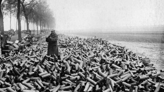 A person in a long coat and hat stands among a massive pile of empty artillery shell casings scattered on the ground beside a row of trees on a misty, barren landscape.