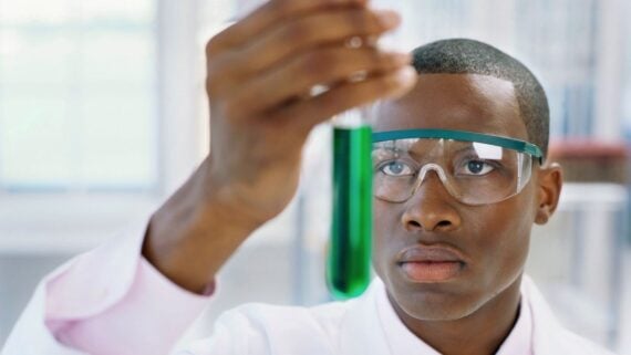 A scientist wearing safety goggles closely examines a test tube containing a green liquid in a laboratory setting.