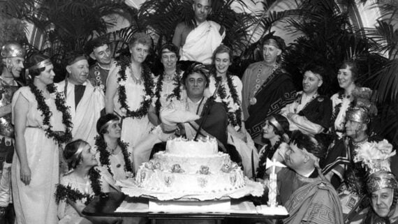 A group of people in Roman-style costumes gather around a large decorated cake, some smiling and looking at the man seated in the center, who is holding a cat. Lush plants fill the background.