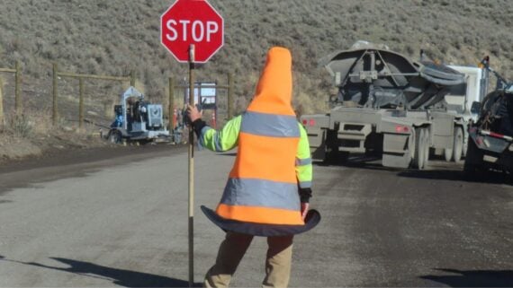 A person wearing a large traffic cone costume and a reflective vest holds a stop sign on a rural dirt road near construction vehicles and equipment.