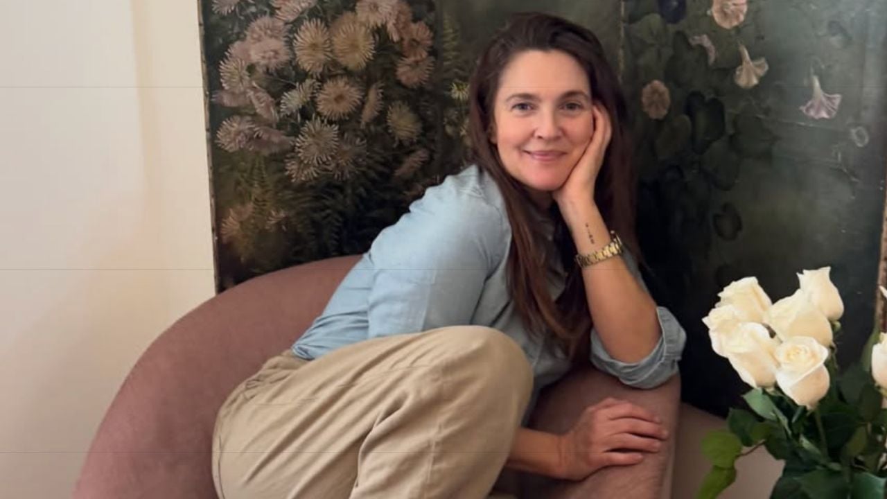 A woman with long brown hair sits casually on a pink chair, smiling and resting her head on her hand. She wears a blue shirt and beige pants. White roses and a floral painting are visible in the background.